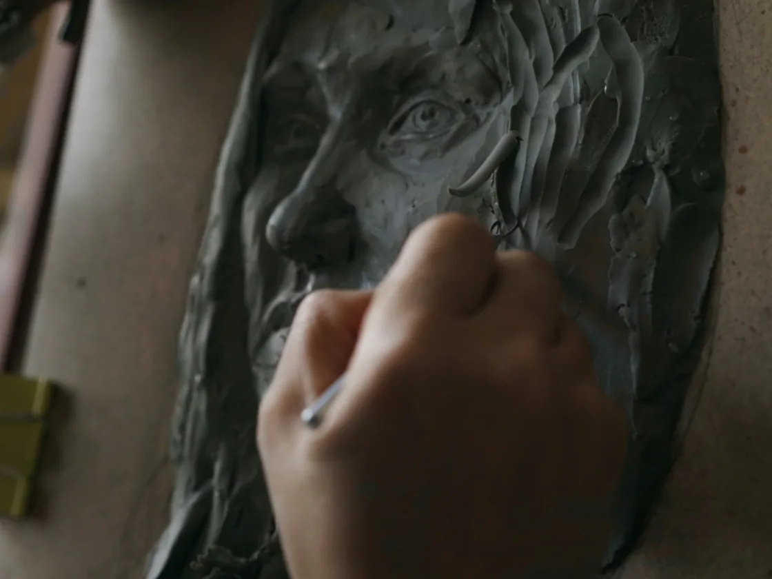 Mano trabajando con un palillo sobre un rostro modelado en barro gris